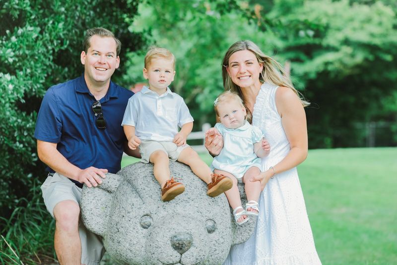 Alaina in a white dress holding her baby daughter, with her husband and their toddler son, gathered around a stone teddy bear in a park