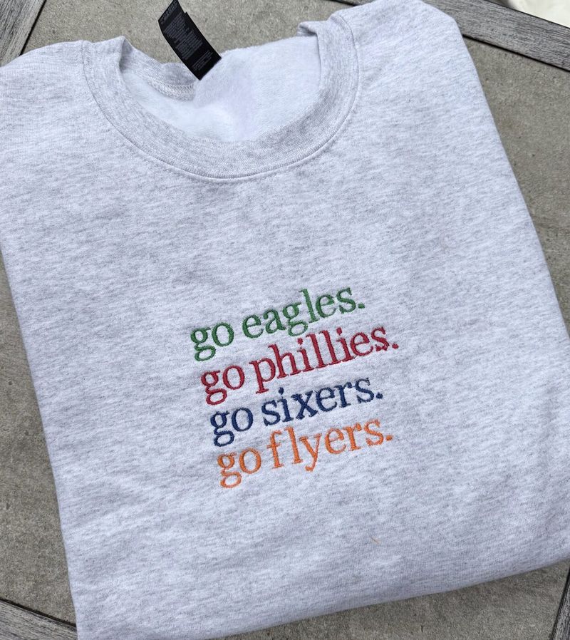 Heather grey crewneck embroidered with four stacked lines: go eagles in green, go phillies in red, go sixers in navy and go flyers in orange