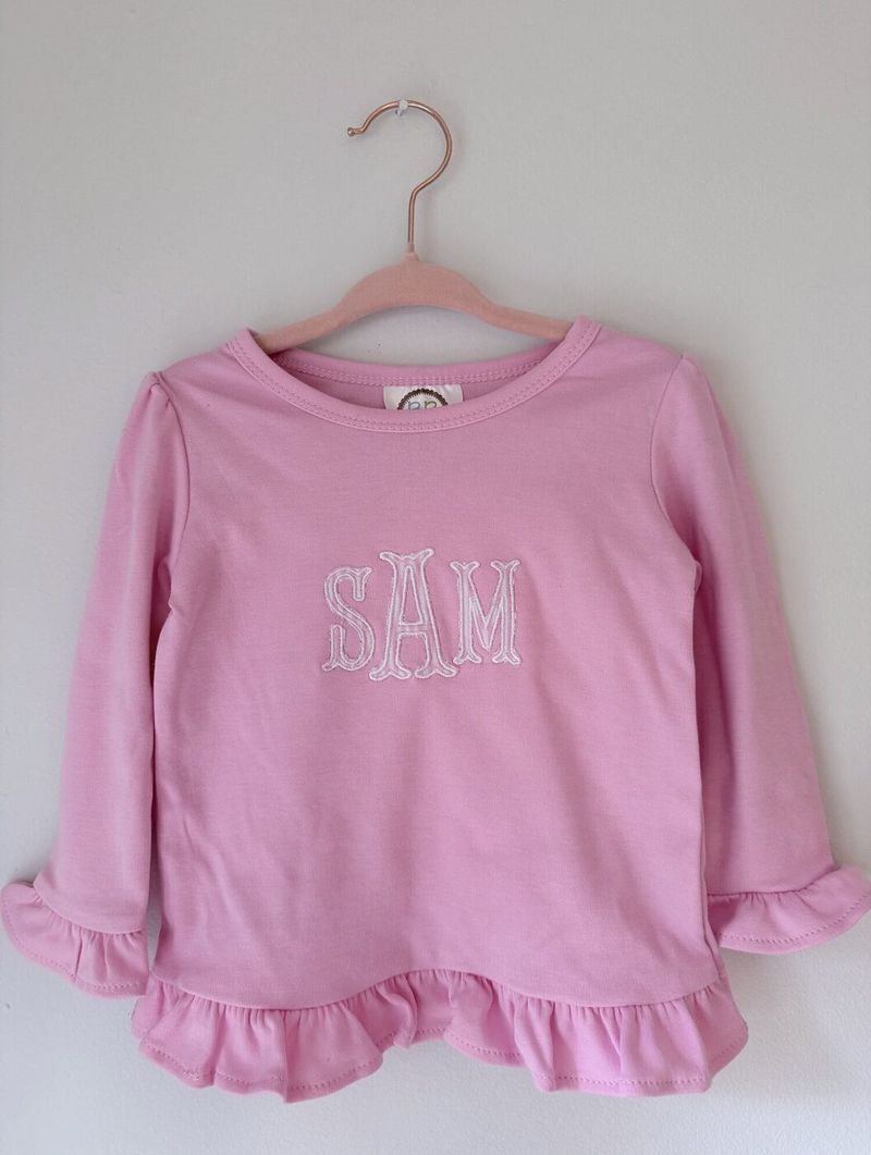 Pink long-sleeve ruffle top with SAM embroidered in white decorative capitals
