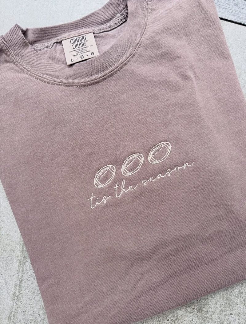 Mauve crewneck with three football outlines above tis the season in script